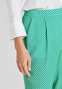 Grüne gemusterte Hose mit einem kreisförmigen Design, elastischem Bund und Falten. Der Stoff hat eine glatte Textur und wird mit einem weißen Oberteil kombiniert.