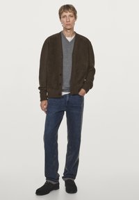 Cardigan marrone con bottoni sopra una maglietta grigia a scollo a V, abbinato a jeans blu e scarpe nere. Tessuto morbido con dettagli a coste vicino ai polsini.