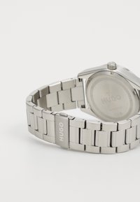 Montre en acier inoxydable argenté avec un bracelet à maillons, présentant un boîtier rond et une boucle de marque gravée "HUGO". Le dos est lisse et gravé.