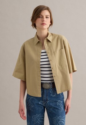 Vrouw draagt een beige korte mouwen blouse met knopen over een wit en zwart gestreept tanktop, blauwe jeans met paisley-patroon en een riem met een ovale gesp.