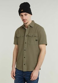Olijfgroene shirt met korte mouwen, twee borstzakken, knoopsluiting en een textuurstof. Gecombineerd met een zwarte beanie met een logo patch.