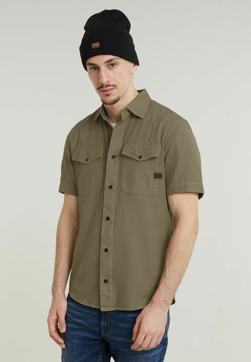 Olijfgroene shirt met korte mouwen, twee borstzakken, knoopsluiting en een textuurstof. Gecombineerd met een zwarte beanie met een logo patch.