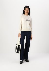 Pull crème avec "voilà" en cursive noire, poignets côtelés, associé à un jean taille haute bleu foncé et des bottines à talons noires. Sac fourre-tout blanc.