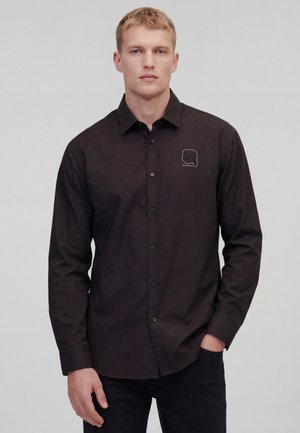 Młody mężczyzna z krótkimi blond włosami, ubrany w czarną koszulę na guziki z małym haftowanym logo na lewej piersi.