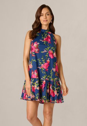 Femme portant une robe bleue sans manches avec des motifs floraux rose vif et vert, debout devant un fond clair uni.