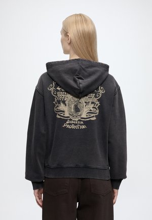 Personne aux longs cheveux blonds porte un sweat à capuche noir avec un graphique ornementé couleur beige et du texte, debout sur un fond clair et uni.