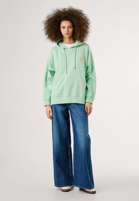 Gaiši zaļa kapuce ar kengūra kabatu, aukliņu kapuci un izšūtu saules detaļu, saskaņota ar platām zilu džinsu biksēm.