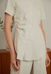 Primo piano di una persona che indossa una camicia a righe chiare con bottoni, caratterizzata da una vestibilità comoda, abbinata a pantaloni coordinati e uno sfondo in legno sfocato.