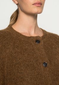 Brun stickad kavaj med rund halsringning, två svarta knappar och en texturerad yta. Mjuk fiber med en mjuk känsla.