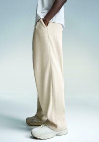 Beige ribbed wide-leg pants with side pockets, paired with light-colored athletic shoes. Background is a gradient from light blue to gray.