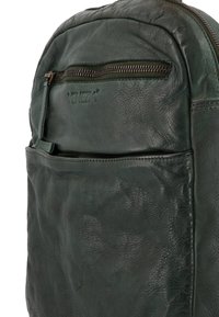 Zelený kožený batoh s texturovaným povrchem, přední kapsou na zip a hřejivým logem. Měkký, poddajný materiál s oblým designem.