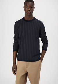 Maglione lavorato a maglia blu navy con motivo a spina di pesce, colletto rotondo e polsini a coste, abbinato a pantaloni beige. Tessuto morbido e testurizzato.
