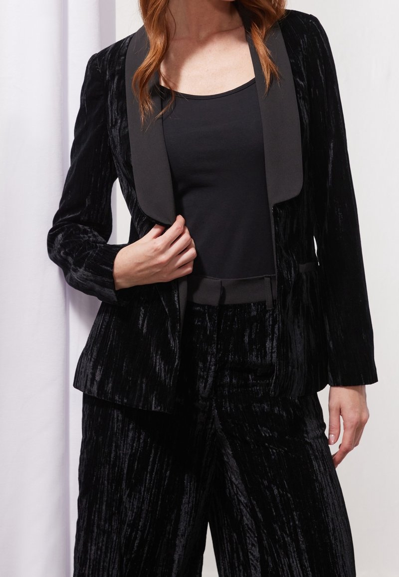 Femme portant un blazer en velours noir avec des revers en satin et un pantalon large en velours assorti, porté sur un top noir près du corps.