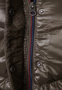 Olivgrön puffjacka med en framträdande dragkedja i röd, blå och svart, tryckknappar och en glänsande, strukturerad yta.