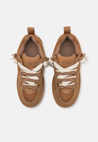 Paire de baskets UGG marron avec lacets blancs et marron, panneaux en mesh texturé et languettes d'accroche sur fond blanc.