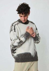 Homme avec des cheveux bouclés portant un pull à motifs noir et blanc, regardant vers le bas et ajustant le poignet avec les deux mains, se tenant devant un fond uni.