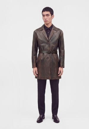 Homme portant un manteau en cuir marron foncé ceinturé par-dessus une chemise noire et un pantalon noir, debout devant un fond clair uni.