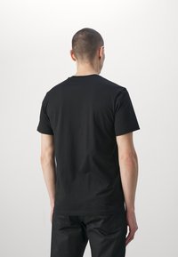 Man med kort hår i en enkel svart t-shirt och svarta byxor, sedd bakifrån mot en vit bakgrund.