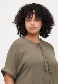 Olive green blouse with short rolled sleeves, a round neckline, and decorative tassels with beads at the front. Smooth fabric texture.