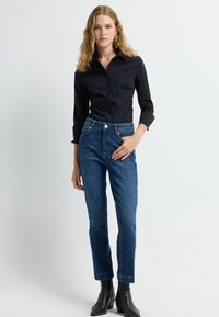 Camisa negra de botones y mangas largas, combinada con jeans de mezclilla azul de tiro alto y botines negros. Diseño limpio, silueta ajustada.