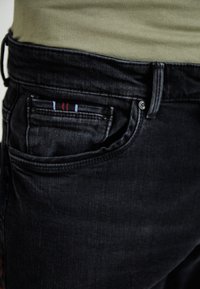 Juodi džinsai su siauru pritaikymu, su priekinėmis kišenėmis, dekoruotomis raudonais, baltais ir mėlynais dryžiais, ir mygtuko užsegimu juosmens srityje.