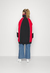 Kvinde med blondt hår i hestehale iført en sort, rød og hvid dunjakke, blå brede jeans og hvide Nike sneakers, set bagfra.