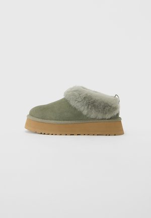 Grønn semsket slip-on sko med tykk brun såle og fluffy lysegrå saueskinnskrage, sett fra siden på en hvit bakgrunn.