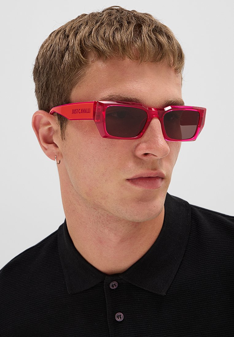 Jeune homme portant des lunettes de soleil rectangulaires rouge vif avec le logo "Just Cavalli" et une chemise noire à col, tourné vers la droite.