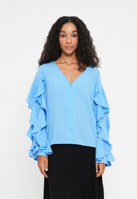Blouse bleu clair avec un col en V, une fermeture à boutons et de longues manches à volants. Le tissu semble lisse et doux, associé à une jupe plissée noire.