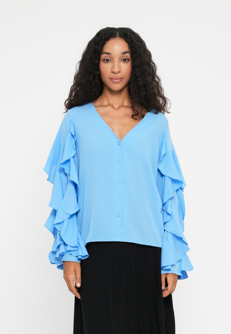Blouse bleu clair avec un col en V, une fermeture à boutons et de longues manches à volants. Le tissu semble lisse et doux, associé à une jupe plissée noire.