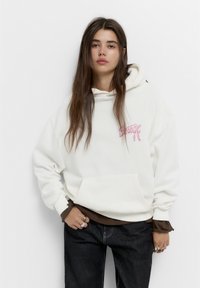 Hvid hættejakke med en frontlomme og pink broderet tekst. Laget over en brun skjorte, parret med mørke jeans, der viser en afslappet pasform.