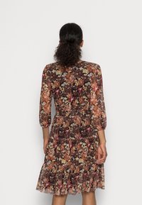 Femme avec des cheveux bouclés attachés, portant une robe marron à motifs floraux, longueur genoux et manches trois-quarts, de dos contre un fond uni.