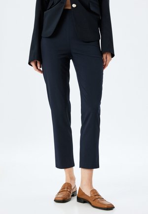 Navy blue tailored pants with a slim fit, cropped length, smooth texture, and minimalistic design. Paired with brown loafers.