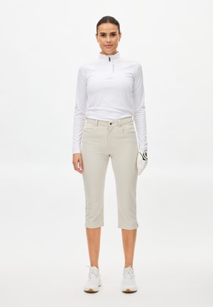 Kobieta stojąca w białej bluzie na zamek z długim rękawem, beżowych spodniach 3/4, białych tenisówkach i białej rękawiczce golfowej na prawej ręce.