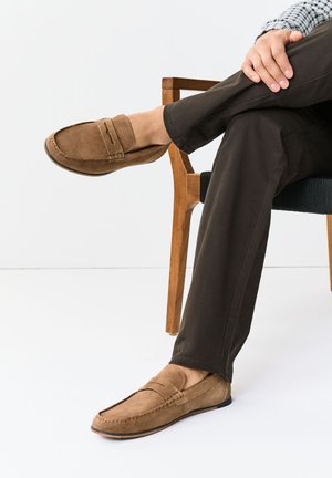 Osoba sediaca so skríženými nohami, oblečená v hnedých semišových mokasínach a tmavých nohaviciach, s rukami položenými na kolene na bielom pozadí.