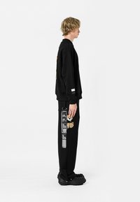 Joven con sudadera negra y pantalones con parches y texto, de perfil contra un fondo blanco liso.