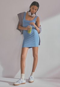 Abito a canotta rigato blu chiaro con taglio dritto. Sneakers bianche, calze crema e maglione grigio appoggiato sulla spalla. Bevanda verde in mano.