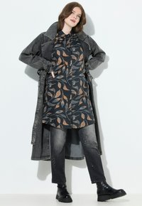 Femme portant une tunique longue à motifs, un jean noir décoloré avec des embellissements, des bottes noires et un long manteau en jean noir délavé avec les mains dans les poches.