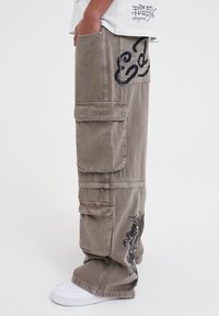 Cargo broek in lichtbruin stof met meerdere zakken. Bevat zwarte grafische ontwerpen en stikdetails. Gedragen met witte sneakers.