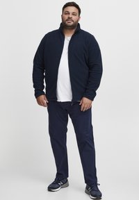 Chaqueta de forro polar azul marino con cremallera, camiseta blanca debajo, combinada con pantalones azul marino y zapatillas deportivas negras. Textura suave, diseño casual.