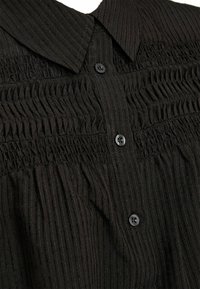 Melnas pogas krekls ar apkakli, kuram ir vertikālas svītras un smockinga detaļas pie pogām, izgatavots no strukturētas auduma.