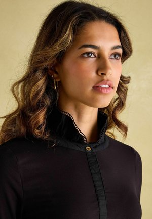 Schwarzes Shirt mit hohem Kragen und Ausschnittdesign, goldener Knopfverschluss und glatter Oberfläche. Das Model hat welliges Haar und trägt Creolen.