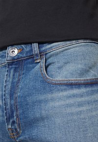 Jeans de mezclilla de color azul claro con cremallera, costuras de cobre y un bolsillo delantero. La cinturilla está parcialmente cubierta por una tela negra.