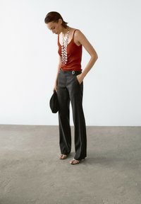 Rotes geripptes Tanktop, schwarze weit geschnittene Hose mit Gürtel und einer silbernen Statement-Halskette. Model trägt transparente Sandalen und hält einen schwarzen Gegenstand.