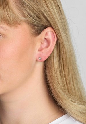 Cercei stud din argint cu design floral, cu pietre prețioase mici și transparente. Prim-plan al unei urechi cu păr lung, blond. Minimalist și elegant.