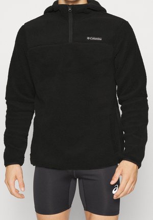 Man draagt een zwarte Columbia fleece-hoodie met een voorrits en zwarte Asics compressieshorts, staat met ontspannen armen.