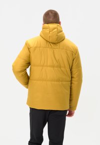 Giacca imbottita gialla con cappuccio, caratterizzata da una texture liscia, cuciture orizzontali e una vestibilità rilassata, indossata sopra pantaloni neri.