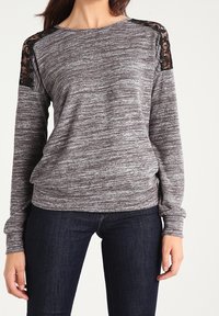 Maglia grigia a maniche lunghe in tessuto testurizzato, con motivo a righe e dettagli in pizzo nero sulle spalle. Abbinata a jeans scuri.