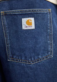 Modrá deninová tkanina so žltým šitím a bielym štítkom s žltým logom a textom "carhartt" na zadnom vrecku.
