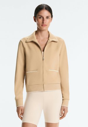 Woman wearing a beige zip-up jacket with front pockets and light beige biker shorts, standing against a plain light background.